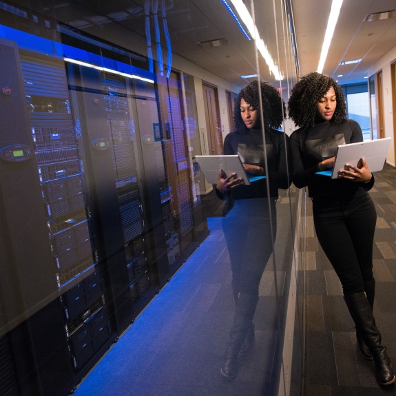 Woman leaning against windows of server room while she is on laptop.